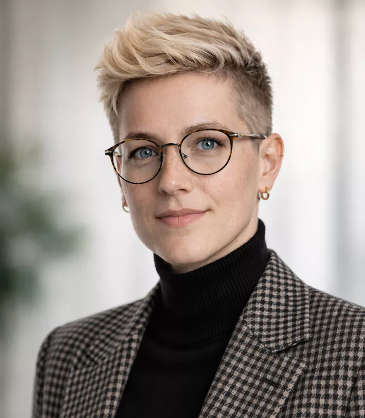 A person with blonde hair is dressed in a checkered blazer and black turtleneck, accessorized with glasses and a gold earring. The setting appears to be indoors with a soft, blurred background. The overall style is modern and sophisticated.